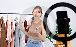 Young Asia woman fashion designer using mobile phone receiving order and showing clothes video live streaming online at shop