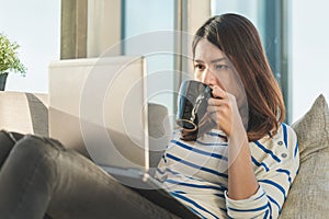 Young asia woman working on a laptop with drinking coffee and sit on sofa at home office