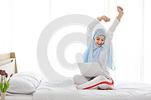 Young asian arab woman using laptop sitting on bed in bedroom. Beautiful muslim girl in sleepwear with blue hijab stretching her