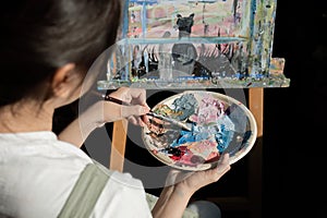 Young Asian artist woman holding a palette of colorful paints is intently painting acrylic paints on canvas in a