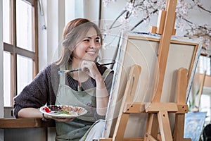 Young Asian artist woman holding a palette of colorful paints is intently painting acrylic paints on canvas in a