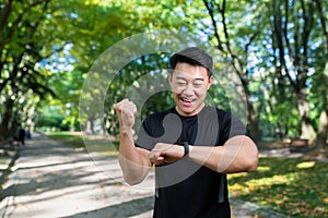 A young Asian athlete man looks at a fitness bracelet, rejoices at the result of a run in the park