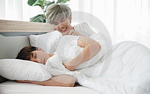 Young Asian attractive gay man smile and provoke his boyfriend by hand put on his shoulder while his lover sleeping on bed in the