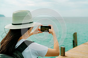 Young Asian backpacker woman wear hat use smartphone taking photo at pier. Summer vacation at tropical paradise beach. Happy