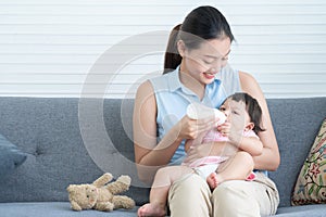 Young Asian beautiful mother smiling, holding and feeding milk from bottle to cute little Caucasian 7 months newborn baby,