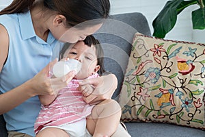 Young Asian beautiful mother smiling, holding and feeding milk from bottle to cute little Caucasian 7 months newborn baby,