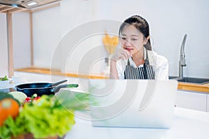 Young asian beautiful woman using a tablet for looking for recipe or learning cooking online with vegetables in kitchen at home,