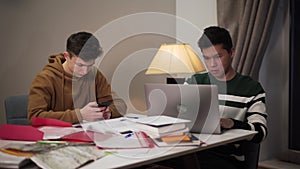Young Asian boy sitting at the table and using laptop as his Caucasian groupmate using smartphone. Two concentrated