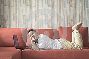 Young asian boy using laptop computer lying on a sofa at home while looking at camera.