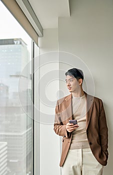 Young Asian business man holding phone looking at window standing in office.