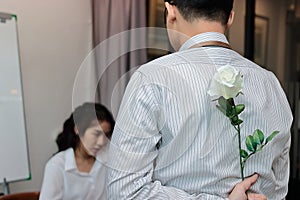 Young Asian business man holding a white rose behind his back for surprise girlfriend in valentines day. Love and romance in offic