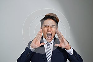 Young Asian business man shouting with hands cupped to his mouth, isolated on gray background
