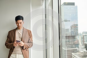 Young Asian business man using mobile phone standing in office at window.