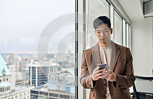 Young Asian business man using mobile phone standing in office at window.