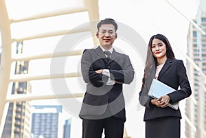 Young Asian business man and woman wearing black suits, standing outdoor with city buildings background, a man crossing his arms