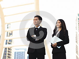Young Asian business man and woman wearing black suits, standing outdoor with light building background, a man crossing his arms