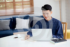 Young asian business man writing on notebook for planning working and using laptop computer on desk at home.