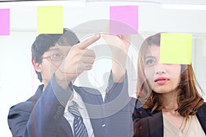 Young Asian business people use post it notes on glass wall to share idea at meeting room. Teamwork and brainstorm concept.