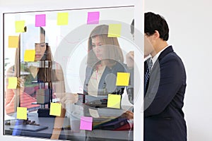 Young Asian business people use post it notes on glass wall to share idea at meeting room. Teamwork and brainstorm concept