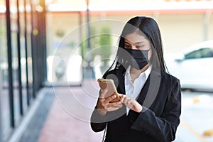 Young asian business woman in business black suit with protect mask for healthcare walking on public outdoor and using smartphone