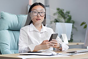 A young Asian business woman in glasses is holding a phone, dialing a message. Sitting in an office