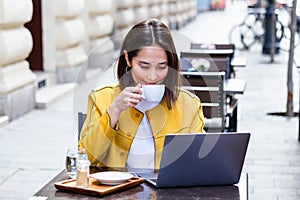 Young asian business woman sitting at table and taking notes in notebook. On table is laptop, smartphone and cup of coffee.On