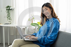 Young asian business woman smile and work from home with laptop computer online to internet on sofa in living room