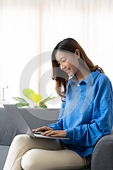 Young asian business woman smile and work from home with laptop computer online to internet on sofa in living room