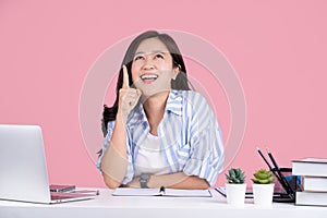 Young asian business woman wear casual shirt. She point index finger overhead on workspace area mock up isolated on pastel pink