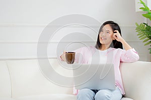 Young asian business woman work from home with laptop computer and drinking coffee on sofa in living room.