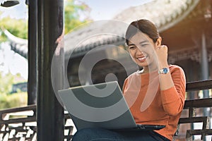 Young asian business woman working with his laptop and enjoying his online winner success on a bench in the park outdoors on