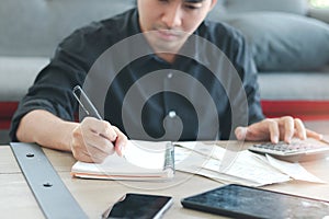 Young Asian businessman in black shirt working on office desk, taking notes while calculate his bill using a calculator, writing