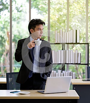 Young asian businessman in black suit drinking coffee while looked outside and thinking of something