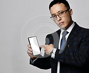 Young asian businessman expert analyst in official jacket, tie and glasses points finger at blank smartphone screen