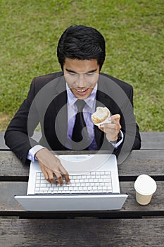 Young Asian businessman having a light snack in a park with his laptop. Conceptual image