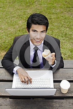 Young Asian businessman having a light snack in a park with his laptop. Conceptual image