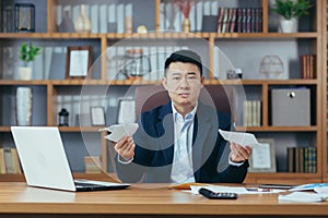 Young asian businessman man, banker, director sitting at desk in office, holding bills, documents, embarrassed, serious, waving