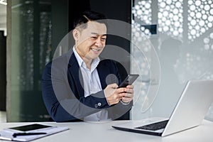 A young Asian businessman sits in the office at the table, holds the phone in his hands, types a message, reads the news