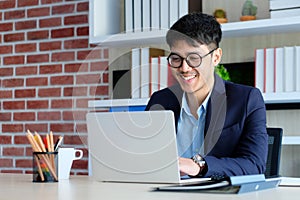 Young asian businessman smiling while working with laptop computer at office, business office lifestyle concept