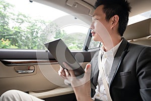 Young asian businessman using tablet and looking out of window while sitting on the back seat in the car.