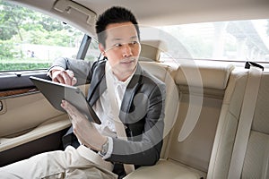 Young asian businessman using tablet and looking out of window while sitting on the back seat in the car.