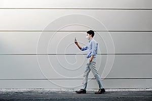 Young Asian Businessman Wearing a Surgical Mask and Using a Smart Phone while Walking by the Urban Building Wall