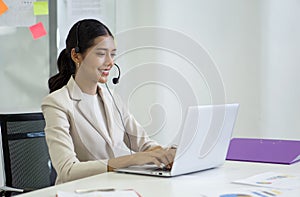 Young asian businesswoman in brown suit and headset with mic typing on laptop computer keyboard while meeting online through