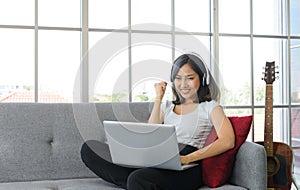 Young asian businesswoman in casual clothing relax wearing headphones in living room at home. punching hand up while working with