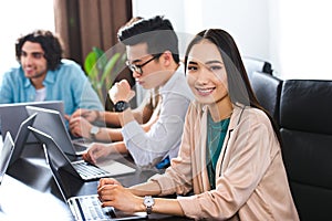 young asian businesswoman looking at camera at table with laptops and businessmen working behind at modern