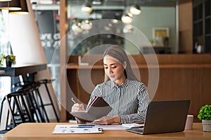 A young Asian businesswoman stands in front of her desk holding a smirked tablet. money business idea