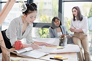 Young asian businesswoman working with blueprint while colleagues sitting behind