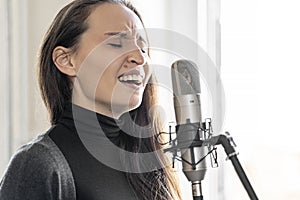 Young Asian or Caucasian woman soulfully singing song to microphone in studio