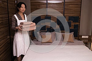 Young asian chambermaid with stack of fresh towels in hotel room