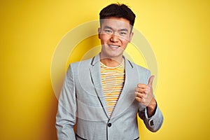 Young asian chinese businessman wearing jacket standing over isolated yellow background doing happy thumbs up gesture with hand